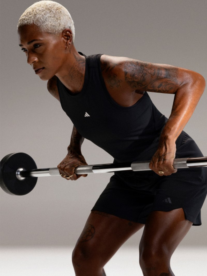 A muscular, tattooed individual in a black tank top is lifting a barbell in a studio setting with a plain background.