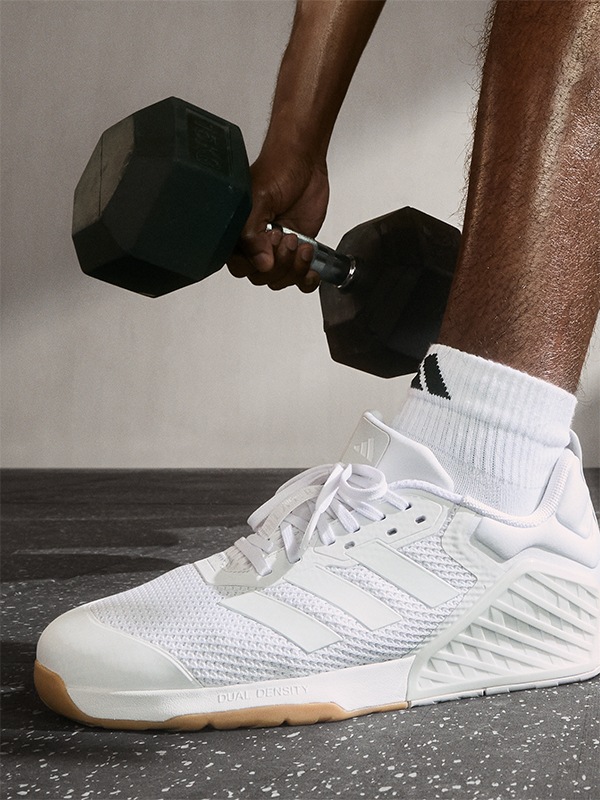 The image shows a person's lower body wearing white sneakers and holding two black dumbbells, against a plain background.