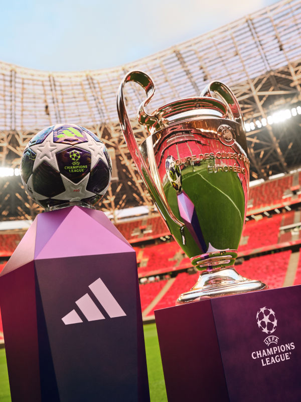 The image shows a UEFA Champions League trophy and a UEFA Champions League match ball on a football stadium field, with the stadium stands visible in the background.