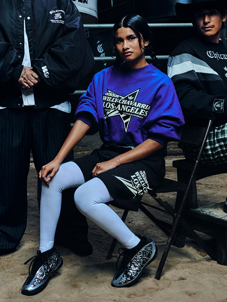 Close-up of a woman wearing a blue adidas x Willy Chavarria 'Los Angeles' top with a large star graphic and Trefoil logo on the front. She stands with her arm resting on the shoulder of a person wearing a black “Chicano” adidas Originals x Willy Chavarria top.