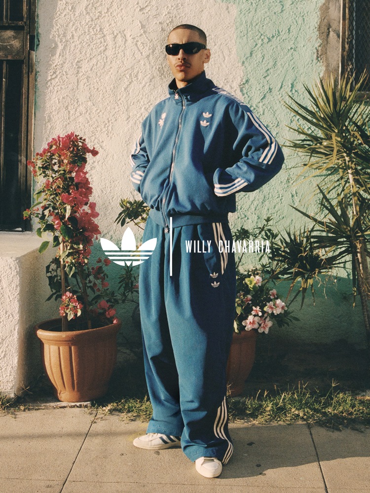 Man with a shaved head stands against a wall surrounded by potted plants. He wears a blue adidas x Willy Chavarria track jacket and matching pants with white stripes and text details. The adidas x Willy Chavarria logo is overlaid on the image.