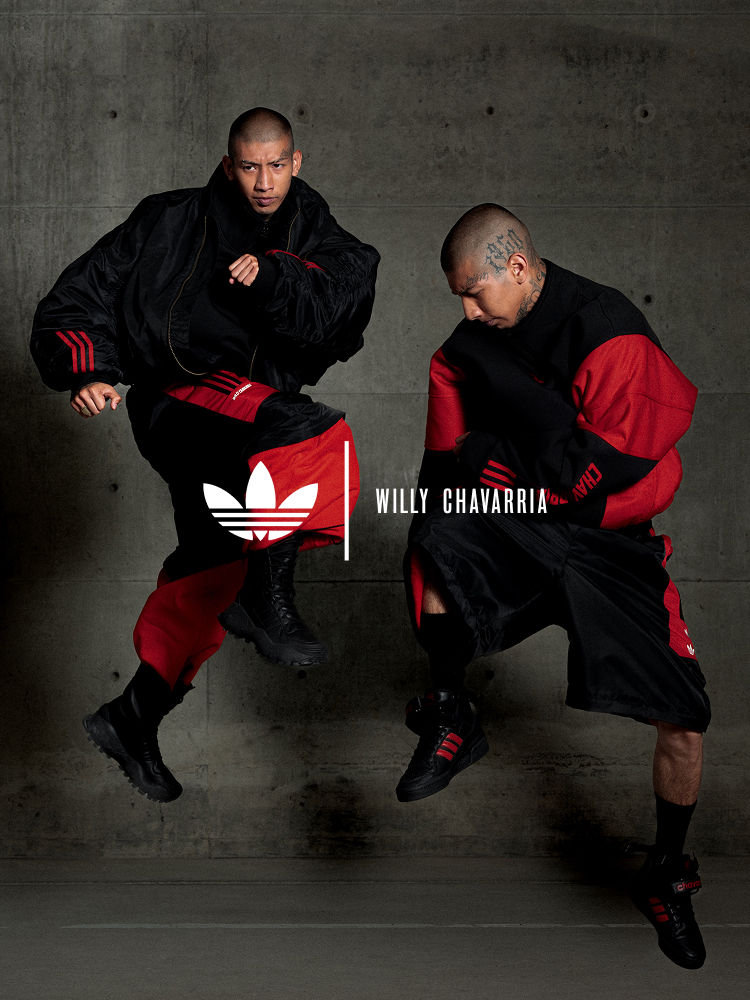 Two men pose in front of a concrete wall wearing black and red outfits from the adidas x Willy Chavarria collection. Both are wearing black adidas sneakers; one pair is the high-top Forum with red stripes, and the other has a chunky, all-black design.
