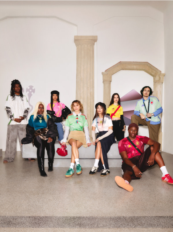 A group of diverse individuals, dressed in vibrant and colorful outfits, are seated together in what appears to be a modern, minimalist interior setting with white walls and columns.
