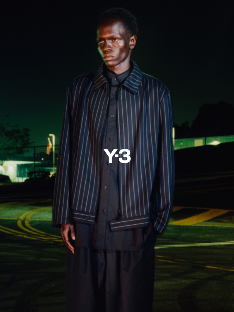 A person wearing a Y-3 branded jacket stands in a dimly lit outdoor setting, with a blurred background suggesting a car park or similar urban environment.