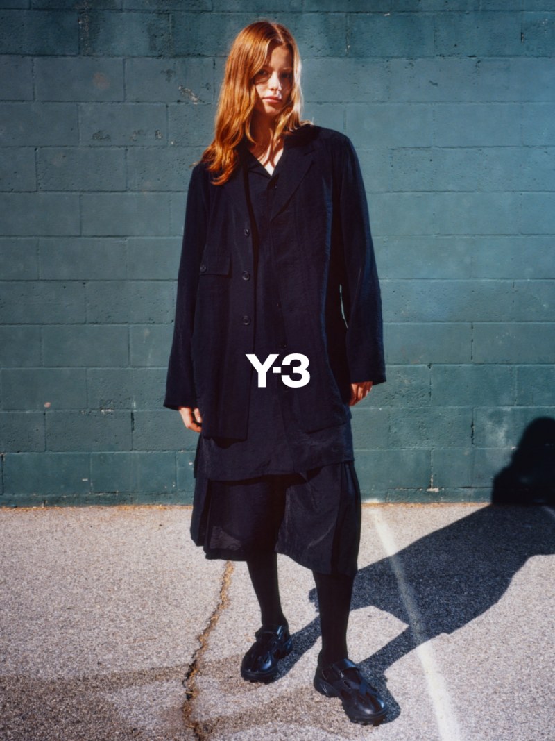 A young woman with long, reddish-brown hair stands in front of a concrete wall, wearing a black Y-3 jacket and matching trousers, and black boots.