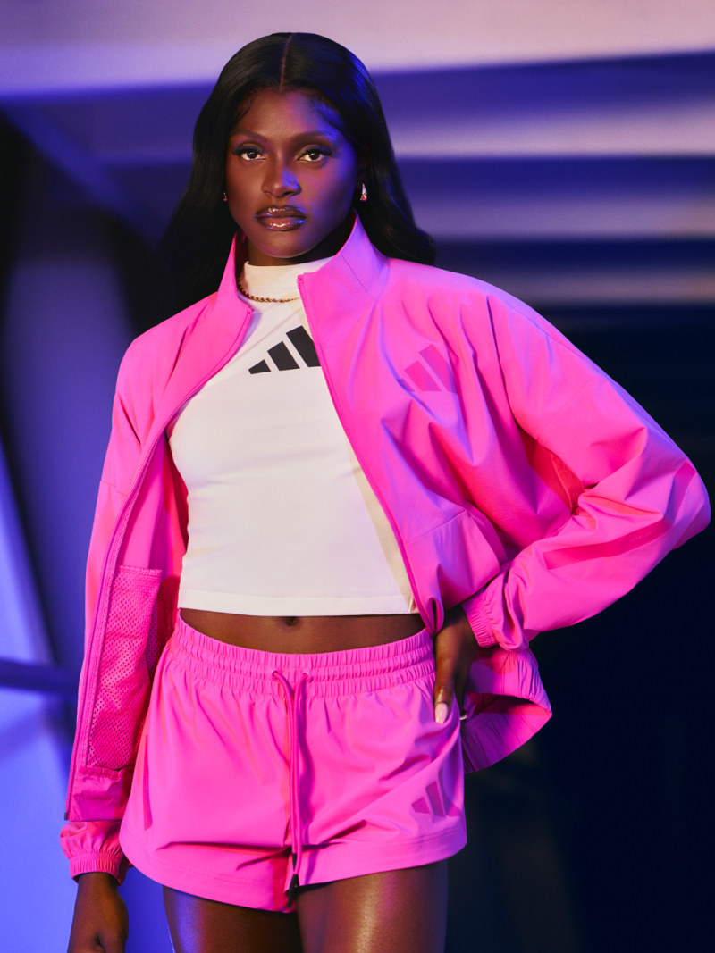 A young woman wearing a bright pink outfit stands against a vibrant purple and blue background, her confident expression and bold fashion choices commanding attention.
