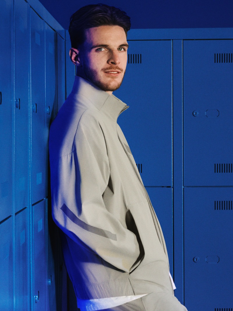 A young man in a light-colored jacket stands in front of blue lockers, his expression serious and focused.