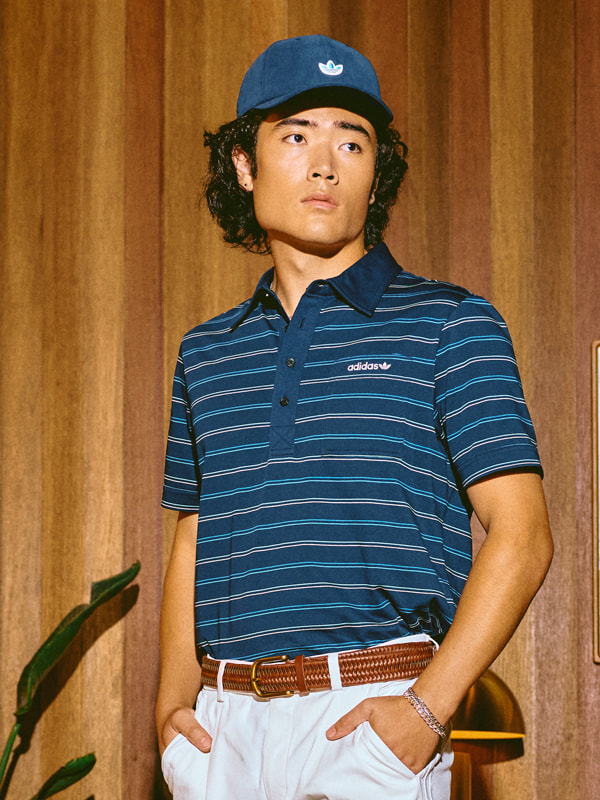 A young man wearing a navy blue striped polo shirt, a navy blue cap, and a brown leather belt stands in front of a wooden background.