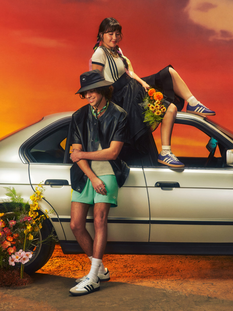 Two individuals, a man and a woman, posing in front of a vintage car against a vibrant, sunset-colored background.