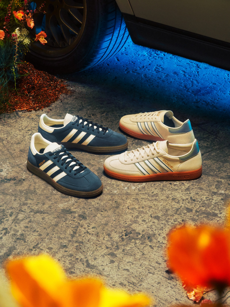 Two pairs of Adidas sneakers, one in navy blue and the other in a beige color, sit on a concrete surface in the foreground, with a colorful holiday wreath and a tire visible in the background.