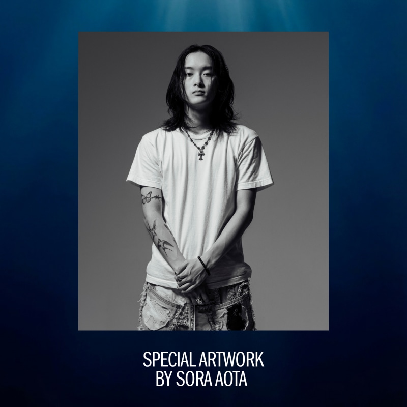 A black and white portrait of a young person with long hair, wearing a white t-shirt and standing against a dark background.