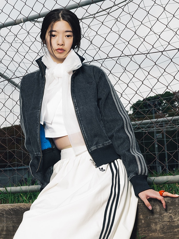 A young woman wearing a black jacket and white scarf stands in front of a chain-link fence, with a grassy field visible in the background.