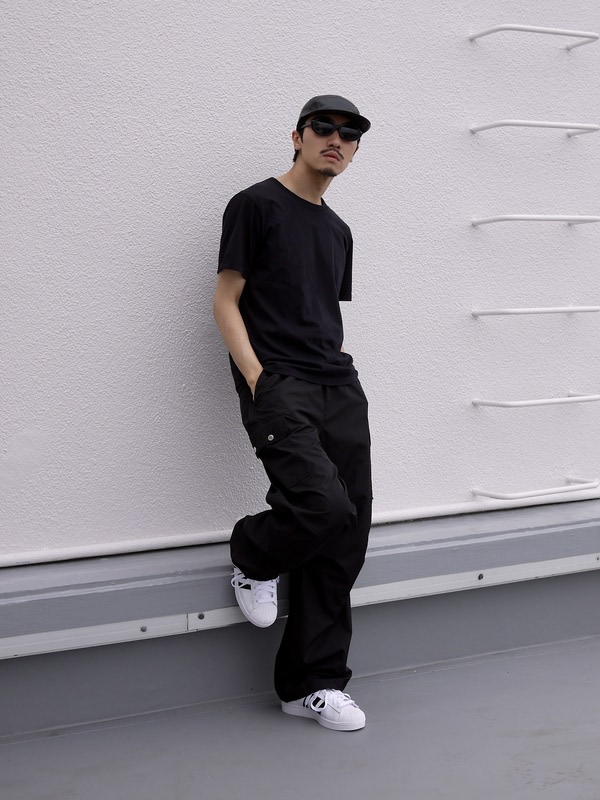 A person wearing a black outfit, including a t-shirt and pants, stands against a gray wall, with white sneakers on their feet.