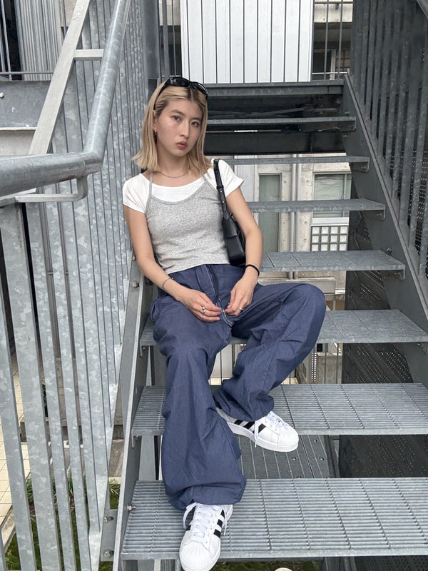 A young woman with blonde hair sits on a metal staircase, wearing a gray top and navy blue pants, with white sneakers on her feet. The background features a gray building with windows.