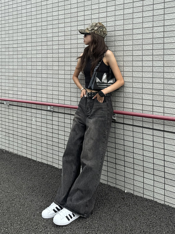 A young woman wearing a black tank top, dark-colored overalls, and a camouflage baseball cap stands in front of a tiled wall.