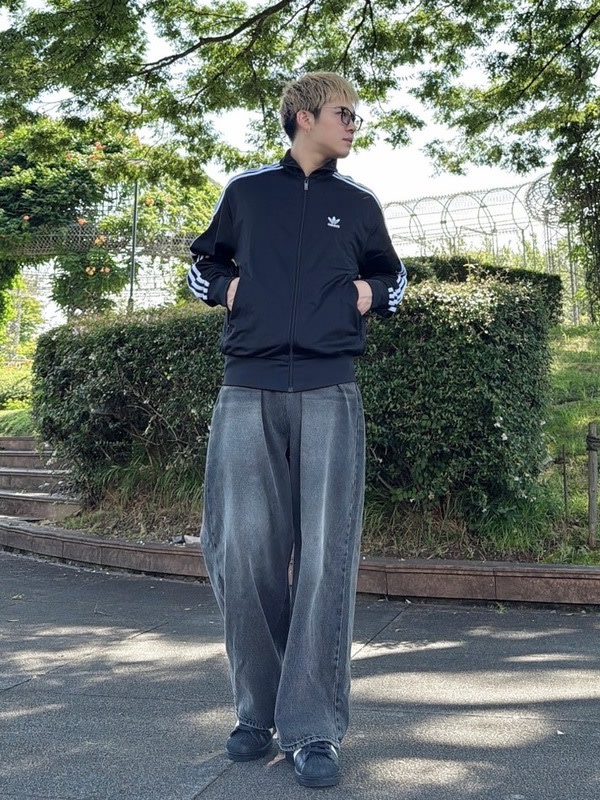 A person wearing a black jacket and gray pants stands in a park-like setting with lush greenery and a fence in the background.
