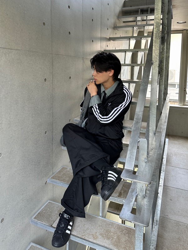 A person wearing a black jacket with white stripes sits on a set of concrete stairs in a minimalist, industrial-style setting.