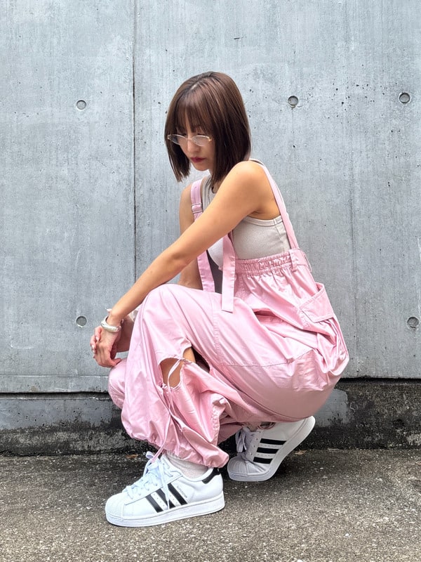 A young girl in a pink dress and white sneakers sits on the ground in front of a concrete wall.