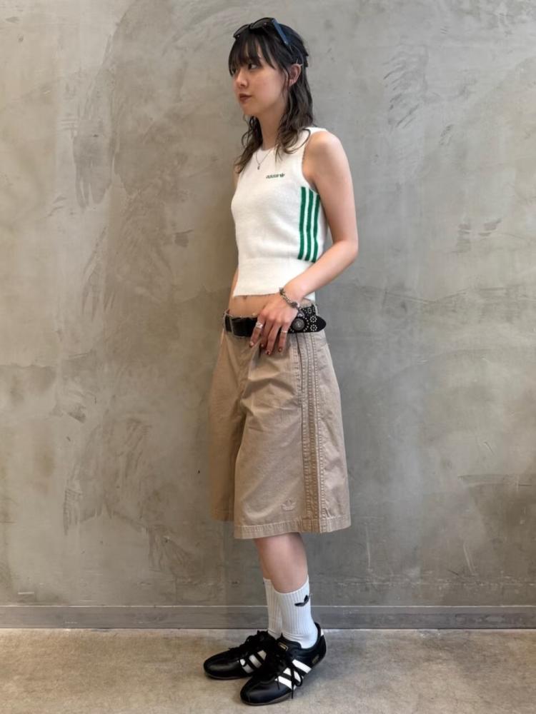 A young woman with dark hair stands against a plain, concrete wall, wearing a white tank top, beige shorts, and black sneakers.