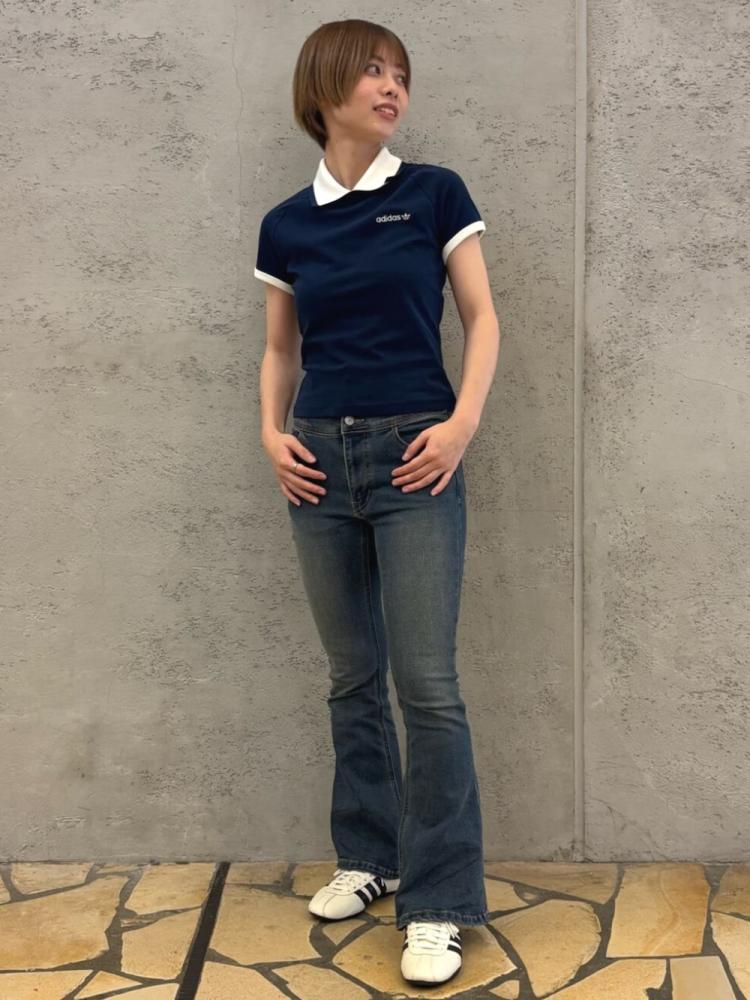 A young woman wearing a navy blue polo shirt and blue jeans stands against a concrete wall, with a paved stone floor visible in the foreground.