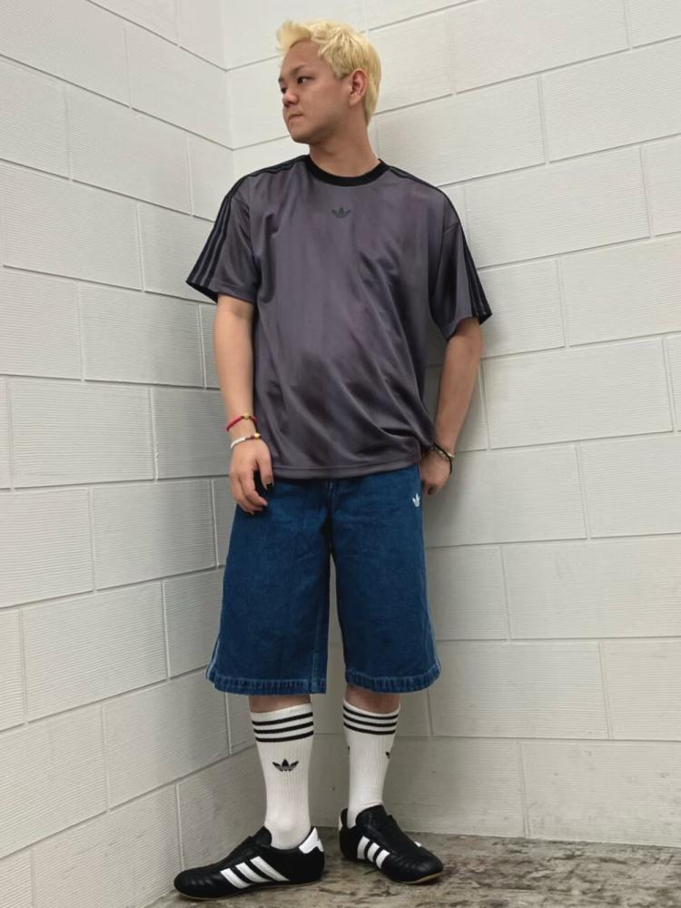A young person with blonde hair wearing a dark shirt, blue shorts, and black and white sneakers stands in a room with white walls.