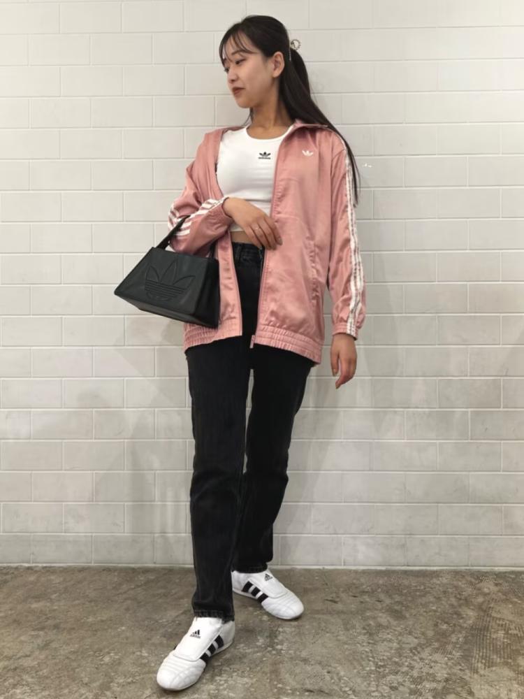 A young woman wearing a pink jacket, white shirt, and black pants stands in front of a plain white wall.