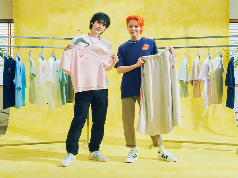 Two individuals, a male and a female, are standing in front of a bright yellow background, surrounded by clothing racks displaying various garments.