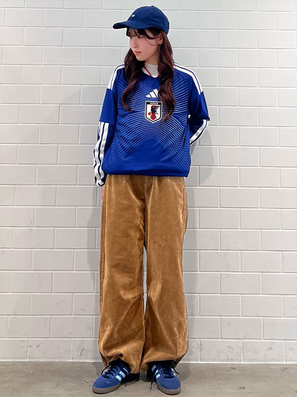 A young woman wearing a blue and white striped jersey, blue baseball cap, and brown corduroy pants stands in front of a white brick wall.