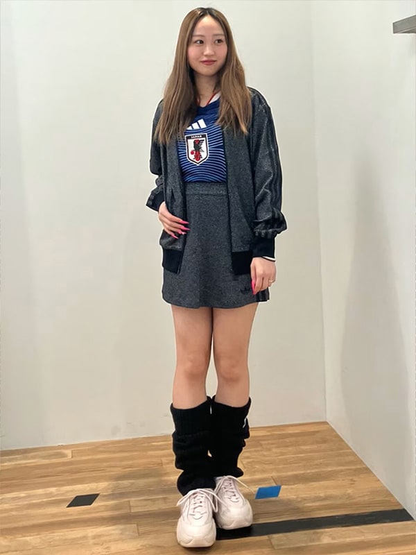 A young woman with long brown hair is standing in a room with a plain white wall, wearing a dark gray jacket, a blue and white jersey, and white sneakers.