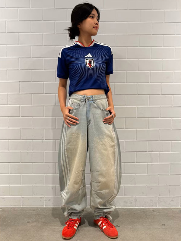 A young person wearing a navy blue sports jersey and loose-fitting gray pants stands in front of a plain white wall.