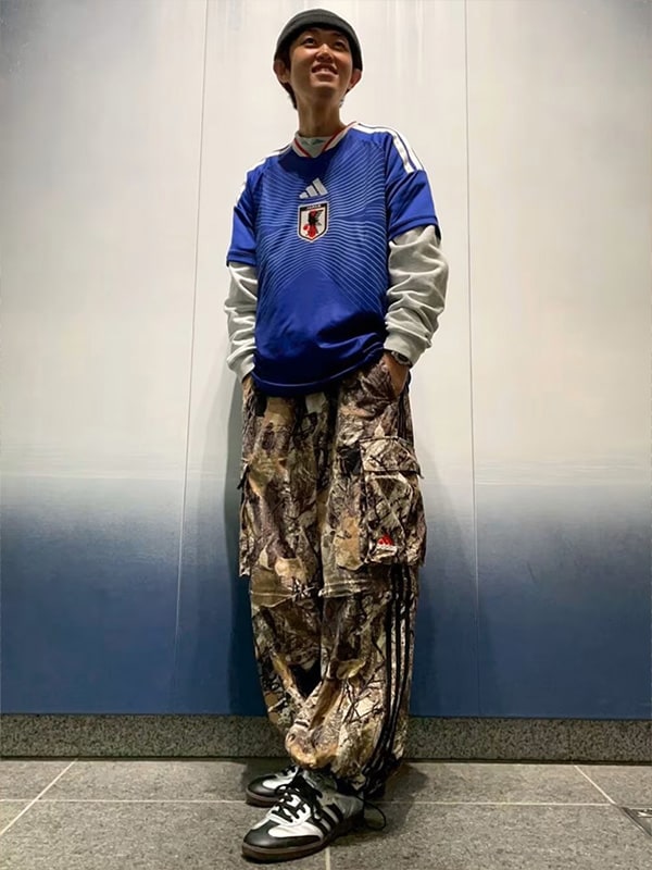 A young person wearing a blue and gray sports jersey, camouflage pants, and black sneakers stands in a plain indoor setting with a blue wall in the background.