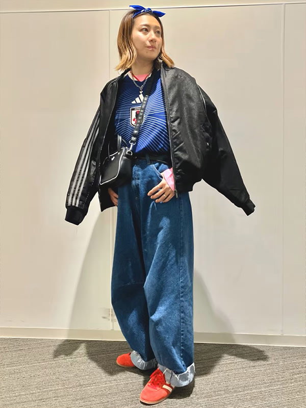 A young woman wearing a black leather jacket, striped top, and loose-fitting jeans stands in a plain, white-walled room.