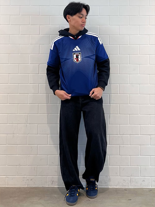 A young man wearing a blue and white soccer jersey stands in front of a white brick wall.