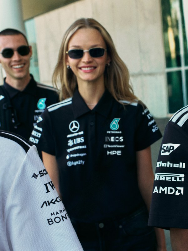 A smiling woman wearing sunglasses and a black racing team uniform stands in front of a racing car.