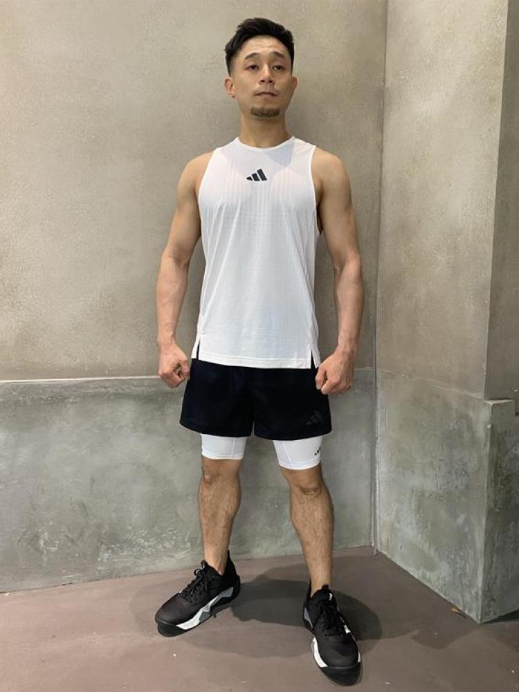 A young man wearing a white tank top and black shorts stands in a plain, concrete-walled room.