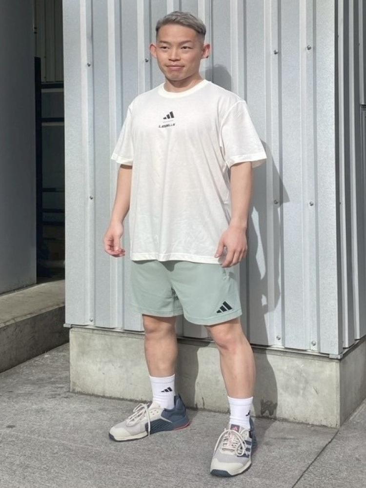A young man wearing a white t-shirt and green shorts stands in front of a gray metal wall, his hands in his pockets and a neutral expression on his face.