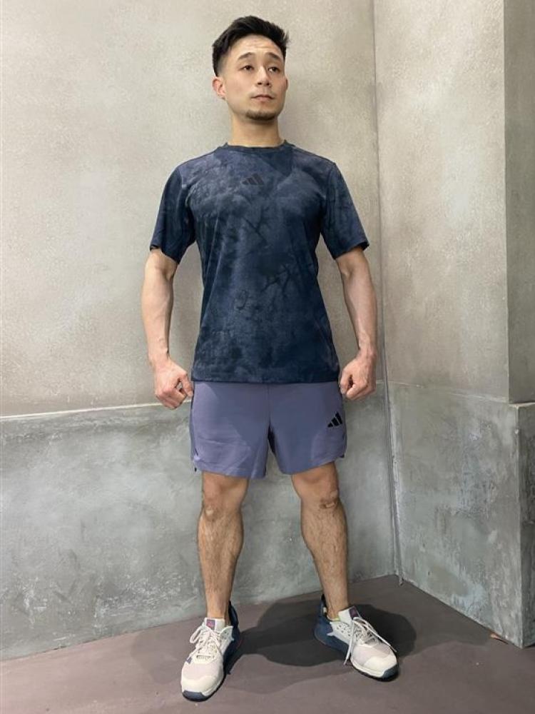 A man wearing a navy blue tie-dye t-shirt and gray shorts stands in front of a plain concrete wall.