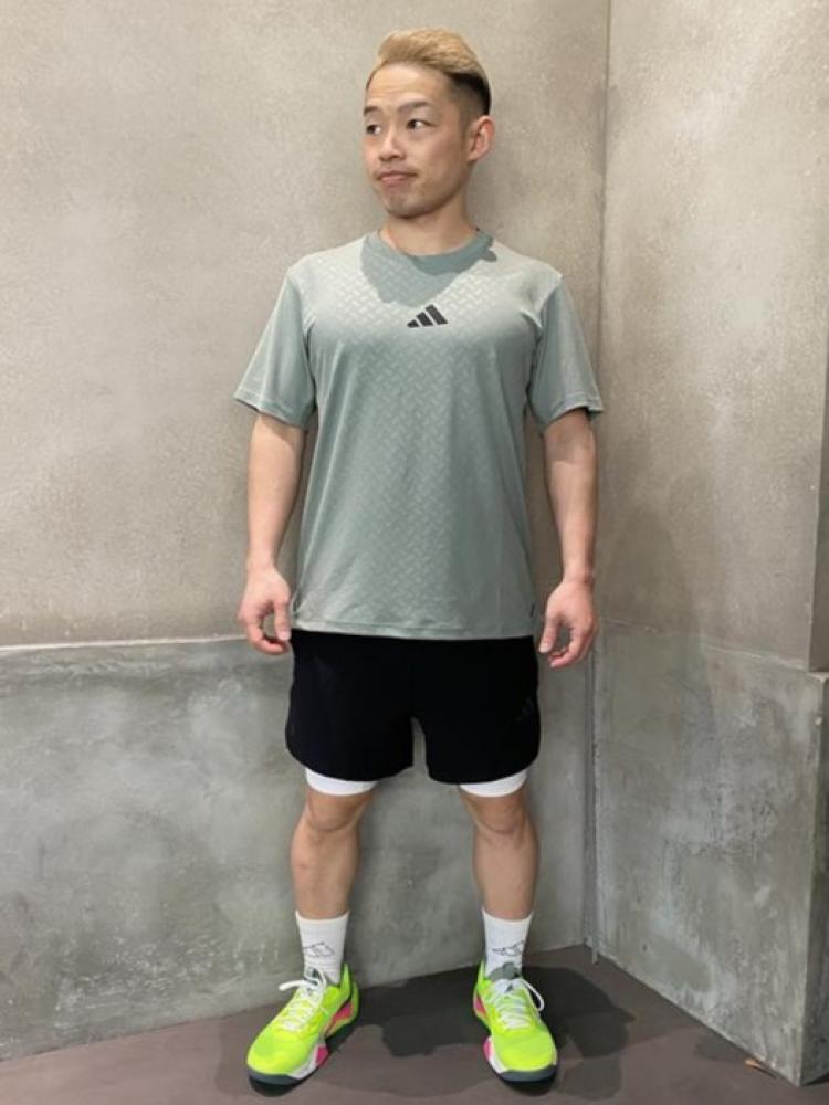 A young man wearing a gray athletic shirt, black shorts, and bright green sneakers stands in front of a plain gray wall.
