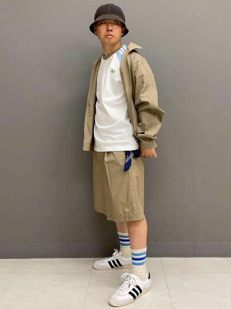 A young person wearing a beige jacket, white shirt, and khaki shorts, standing in front of a gray background.