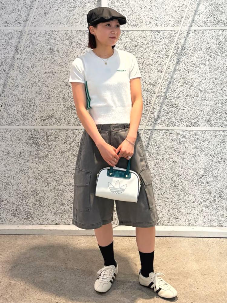 A young woman wearing a white t-shirt, gray shorts, and a baseball cap stands in front of a tiled wall, holding a white handbag.