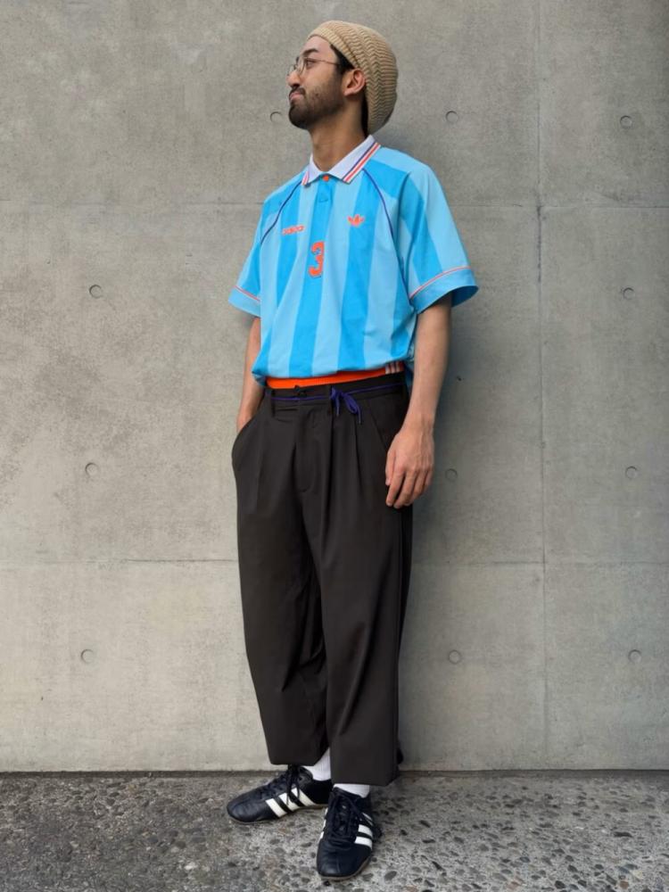 A man wearing a light blue shirt, black pants, and black shoes stands in front of a gray concrete wall.