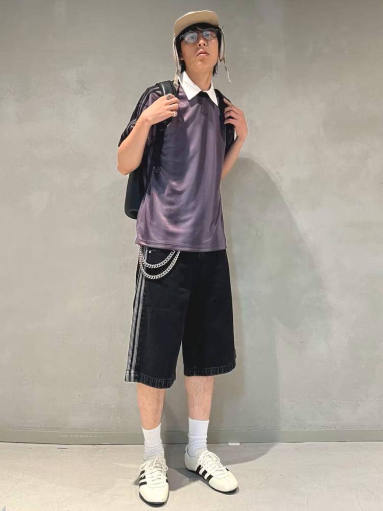 A person wearing a purple shirt, black shorts, and white sneakers stands in front of a plain gray background.
