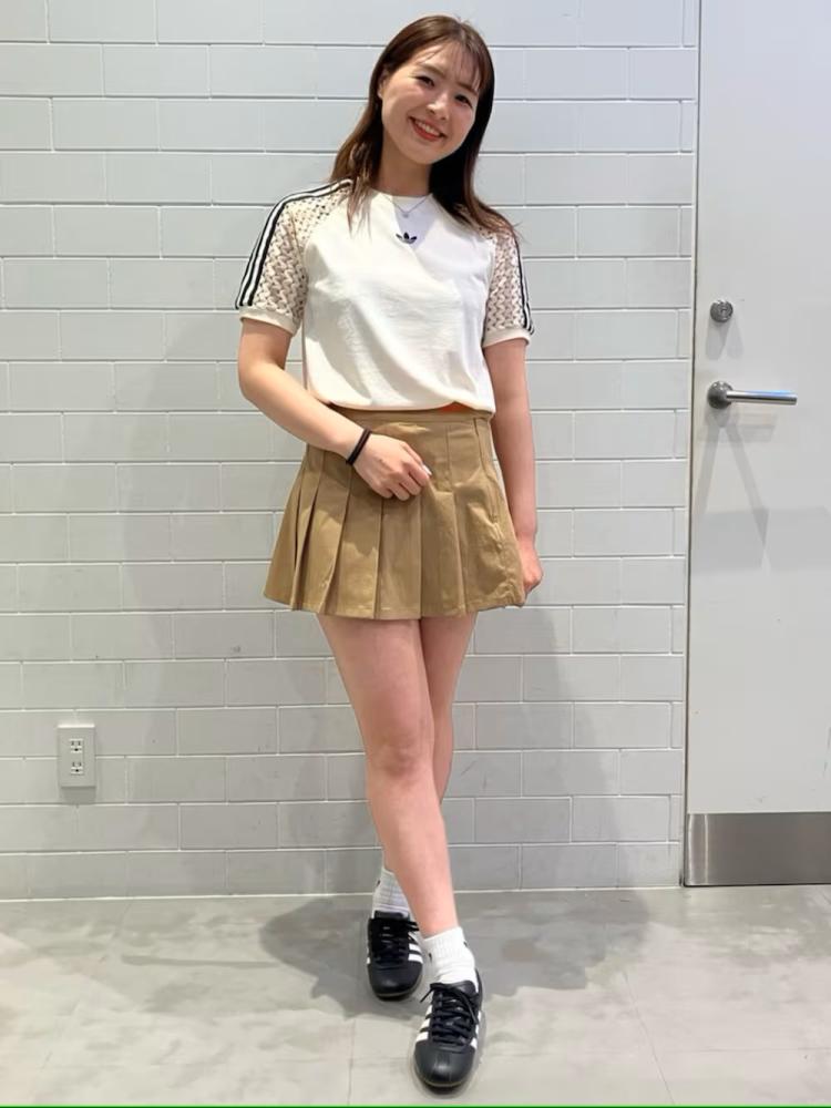 The image shows a young woman with long dark hair wearing a white top and a beige pleated skirt, standing in front of a plain gray wall.
