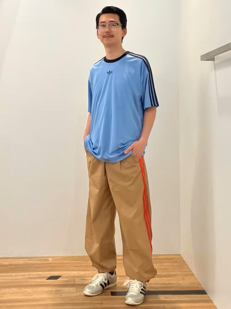 A young man wearing a blue t-shirt and beige pants stands in a plain room with a wooden floor.