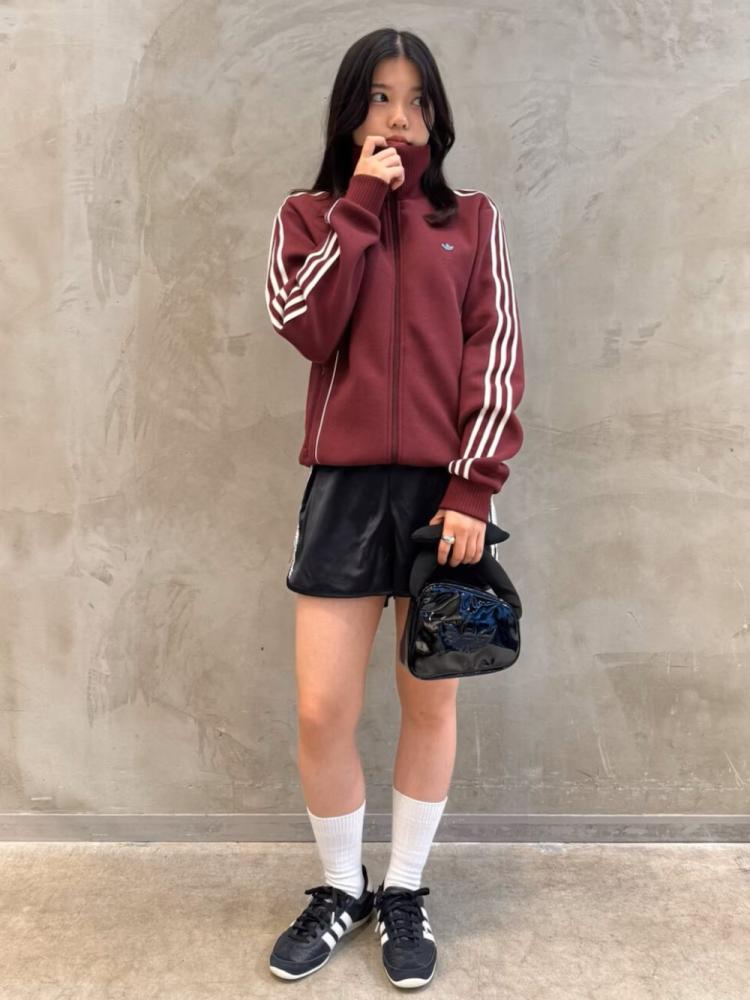 A young woman wearing a burgundy jacket with white stripes, black shorts, and white socks stands in front of a plain gray wall.