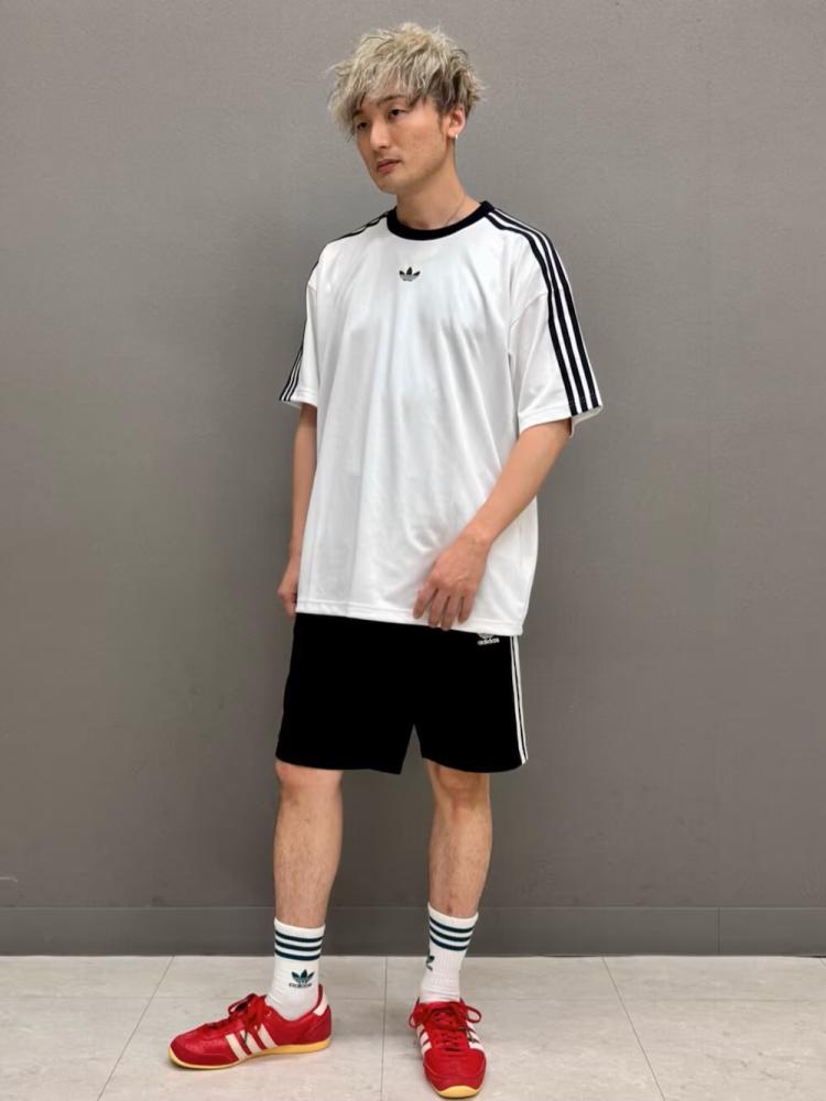 A young person wearing a white Adidas t-shirt, black shorts, and red Adidas sneakers stands against a plain gray background.