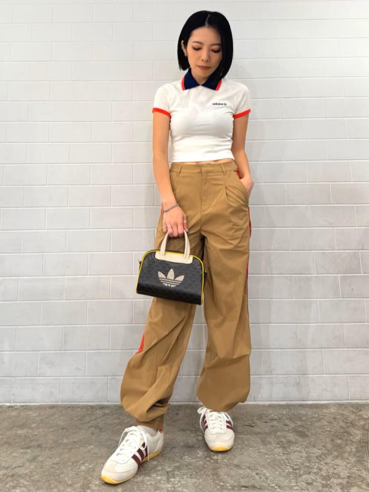 A young woman wearing a white top, beige pants, and white sneakers stands in front of a plain white wall, holding a black and white handbag.
