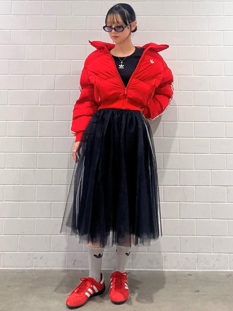 A person wearing a bright red puffy jacket, a black top, a black tulle skirt, and red sneakers stands in front of a plain white wall.