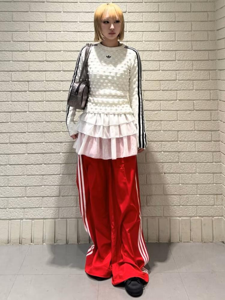 A person wearing a white polka dot top, a ruffled pink skirt, and bright red pants with white stripes, standing in front of a plain white brick wall.