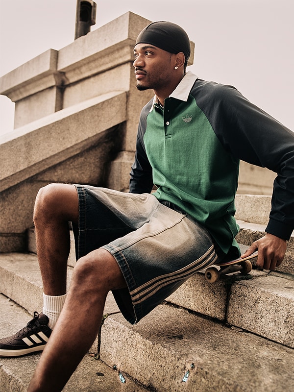 A young man wearing a green polo shirt and black shorts is sitting on a set of stone steps, with a building visible in the background.